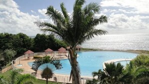 En pool vid ett semesterhotell med en palm i förgrunden, omgiven av avkopplande sittområden och med utsikt över havet.