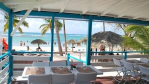 En restaurang vid stranden med uteservering och utsikt över havet, med en byggnad som har ett tak av halmtak och palmer runt omkring.
