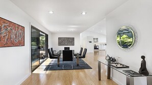 A modern interior with a dining area, kitchen, and a large mirror reflecting greenery. - Arana A Little Blue Mountains Luxury (WENTWORTH FALLS)