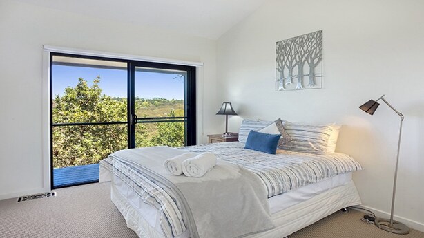 A bedroom with a large window, a bed with white bedding, a nightstand, a lamp, and a wall art piece. - Arana A Little Blue Mountains Luxury (WENTWORTH FALLS)
