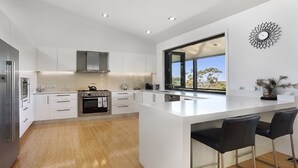A modern kitchen with white cabinetry, a large island, stainless steel appliances, and a view of the outdoors. - Arana A Little Blue Mountains Luxury (WENTWORTH FALLS)