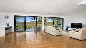 A modern living room with a large sliding glass door leading to a balcony, a sofa, a TV, and a dining area with chairs. - Arana A Little Blue Mountains Luxury (WENTWORTH FALLS)
