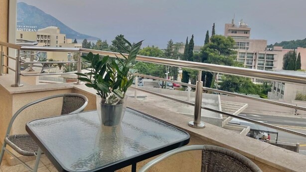 A balcony with a table and chairs, a potted plant, and a view of a cityscape with buildings and a mountain in the distance. - Apartments Boreta Becici (Becici)