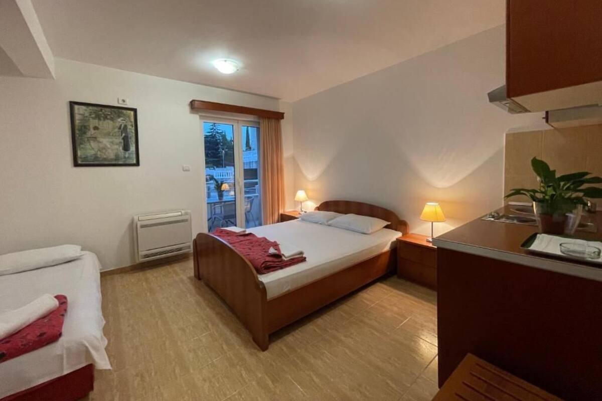 Ein Hotelzimmer mit Bett, Nachttischen, Schreibtisch, Stuhl, einem Fenster mit Vorhängen, Heizkörper und einer Topfpflanze.