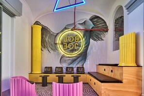 A room with a large mural of an eye and wings, a neon sign reading "JOB", and a reception desk with chairs.