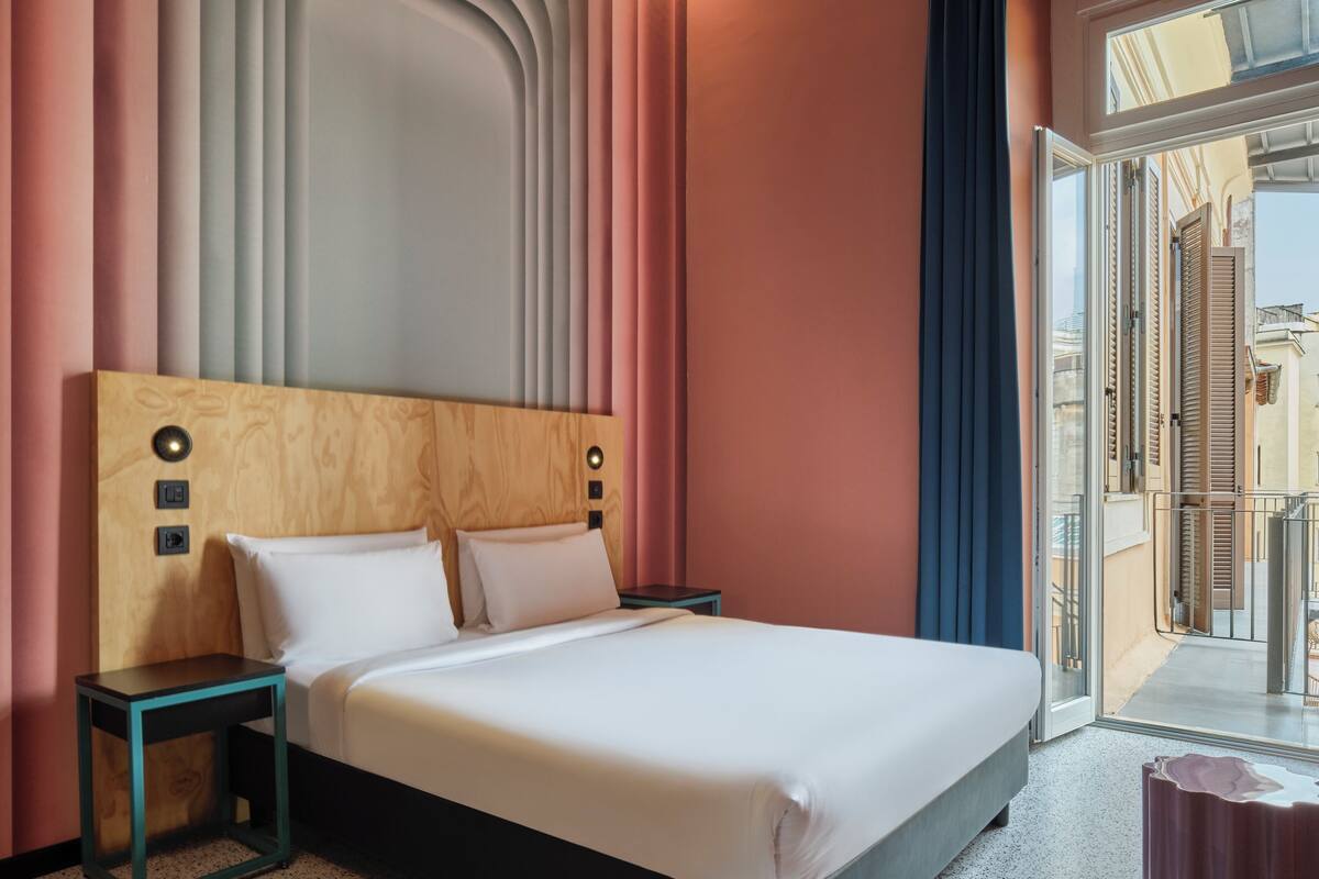 Ein Hotelzimmer mit Holz-Kopfteil, einem Bett mit weißer Bettwäsche, einem Nachttisch und Blick durch eine Glastür ins Freie.