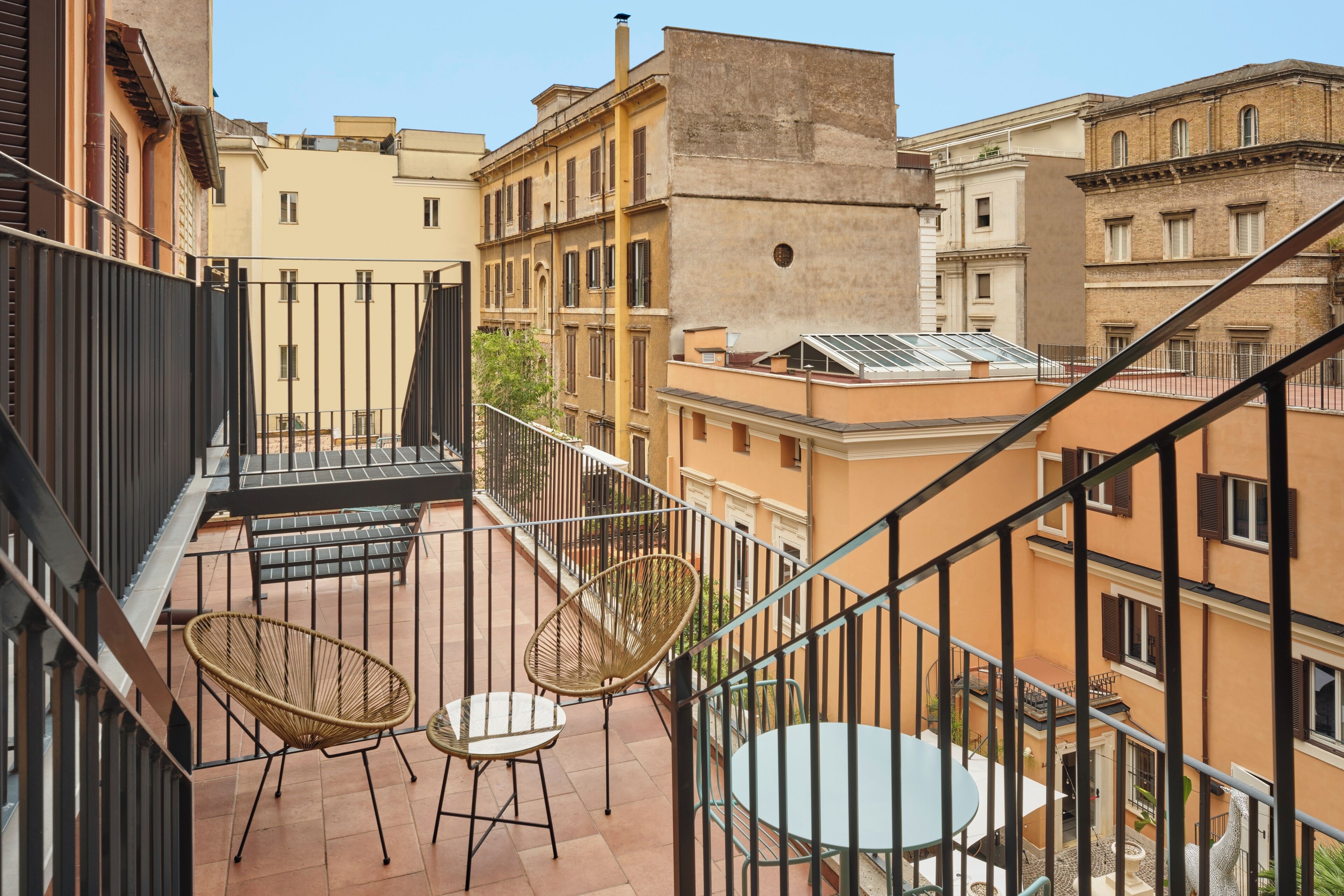 A balcony with wicker chairs and a table, overlooking a cityscape with terracotta buildings.