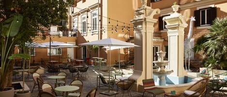 An outdoor cafe with tables and chairs, a fountain, and string lights.
