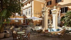An outdoor cafe with tables and chairs, a fountain, and string lights.