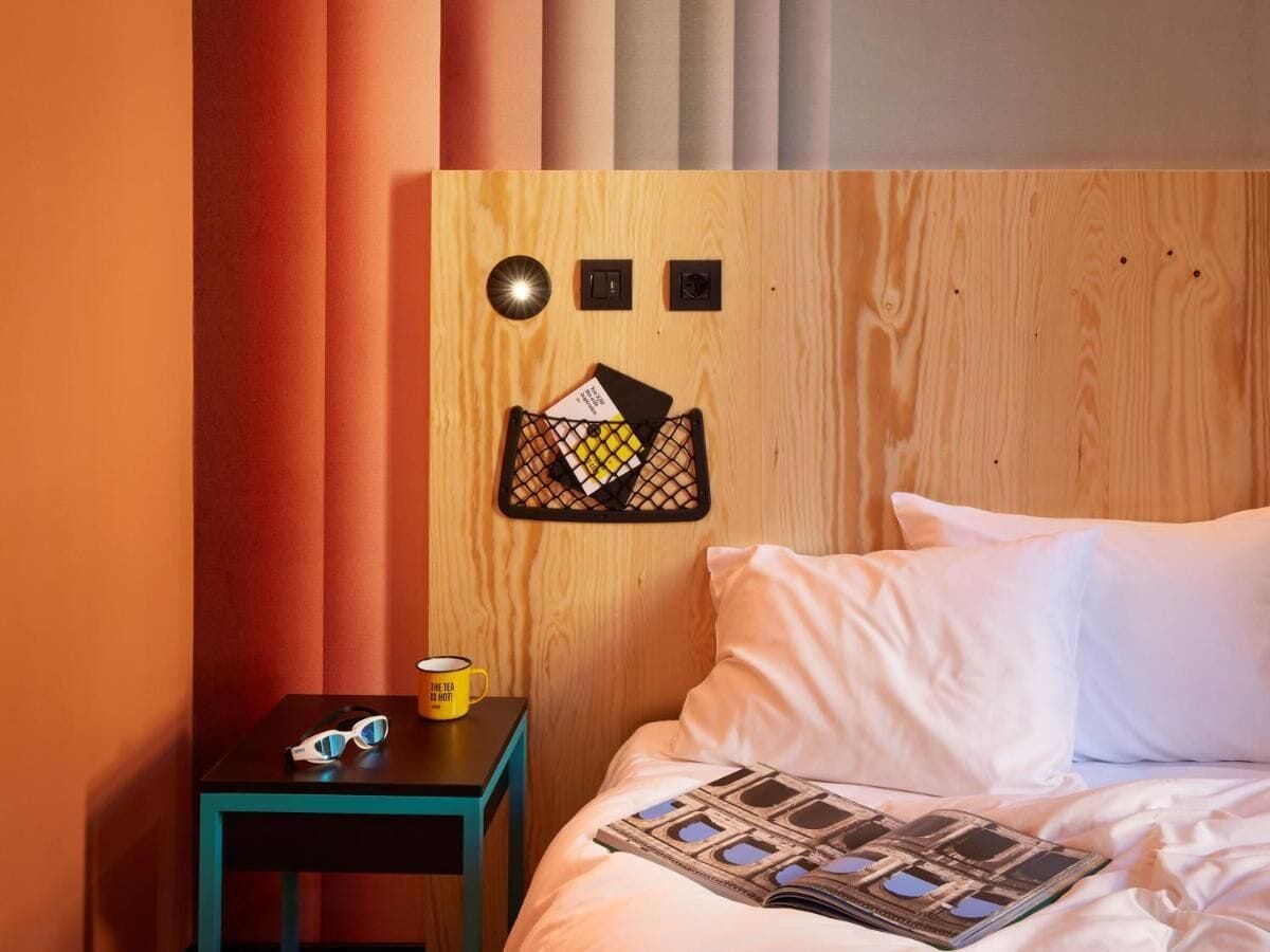 Uma cama bem arrumada com lençóis brancos, uma pequena mesa de cabeceira com uma caneca amarela e um luminário fixado na parede.