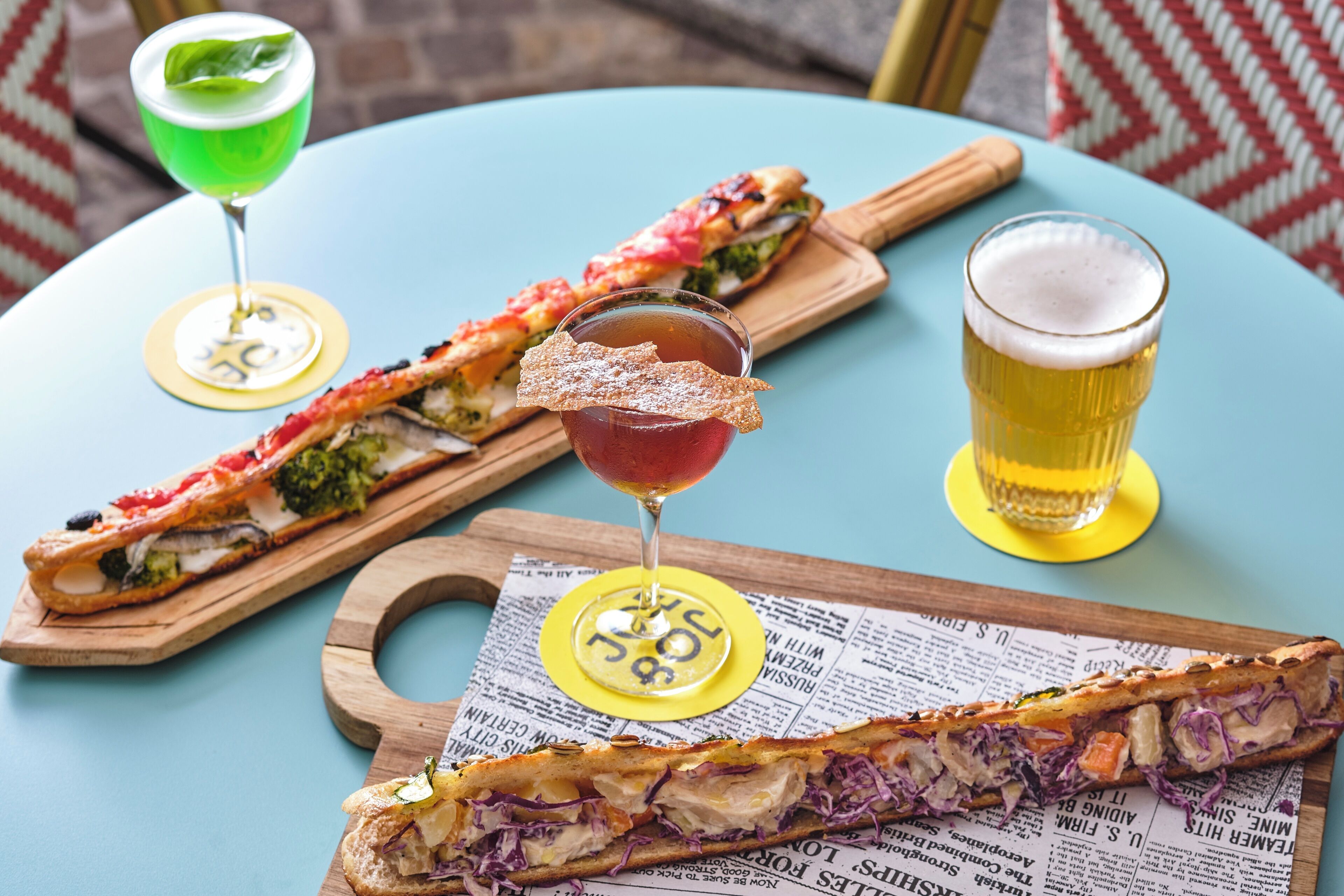 Una mesa con dos sándwiches y dos bebidas sobre posavasos, y un periódico debajo.