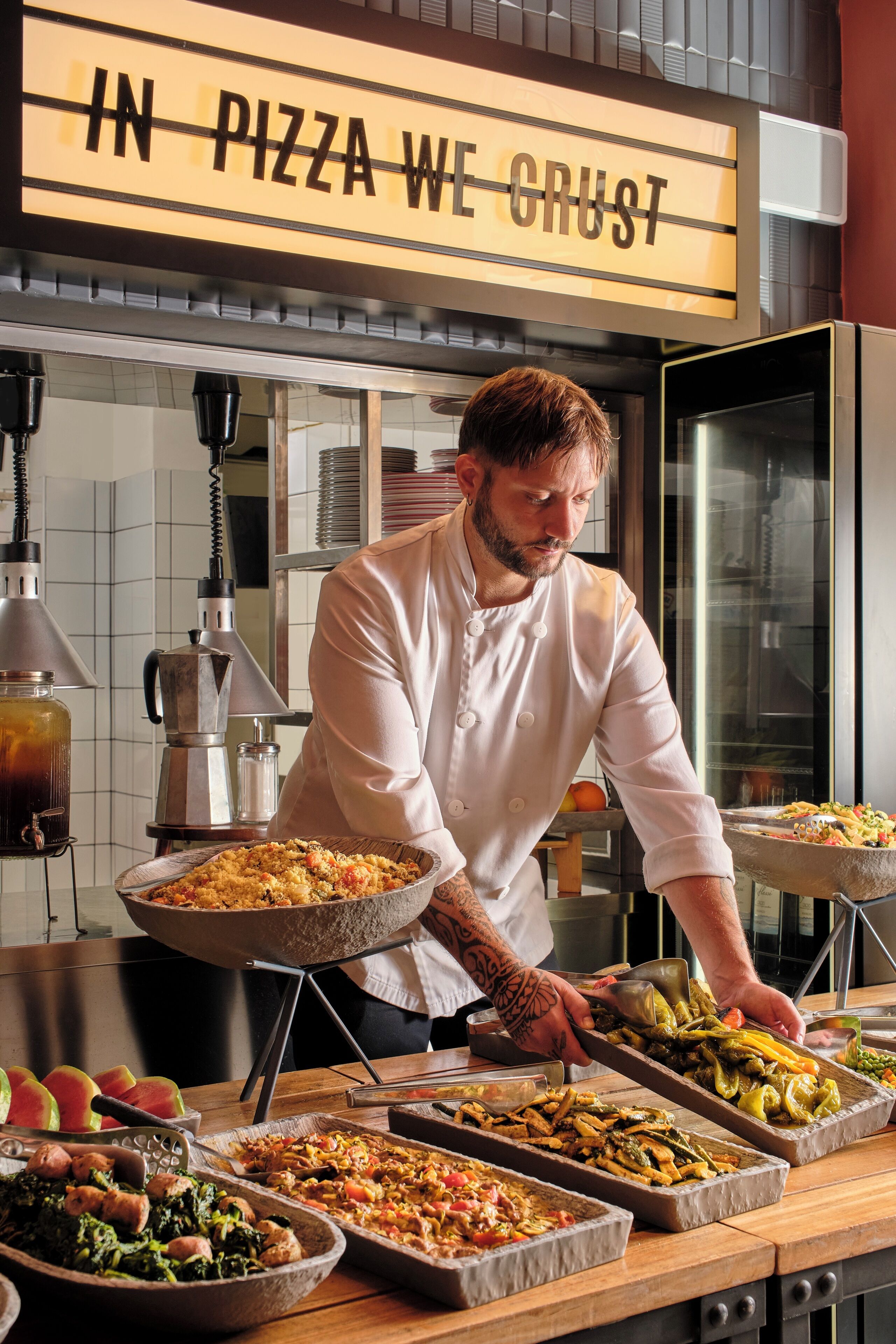 A chef in a white uniform serving food at a buffet with a sign above that reads "IN PIZZA WE CRUST."