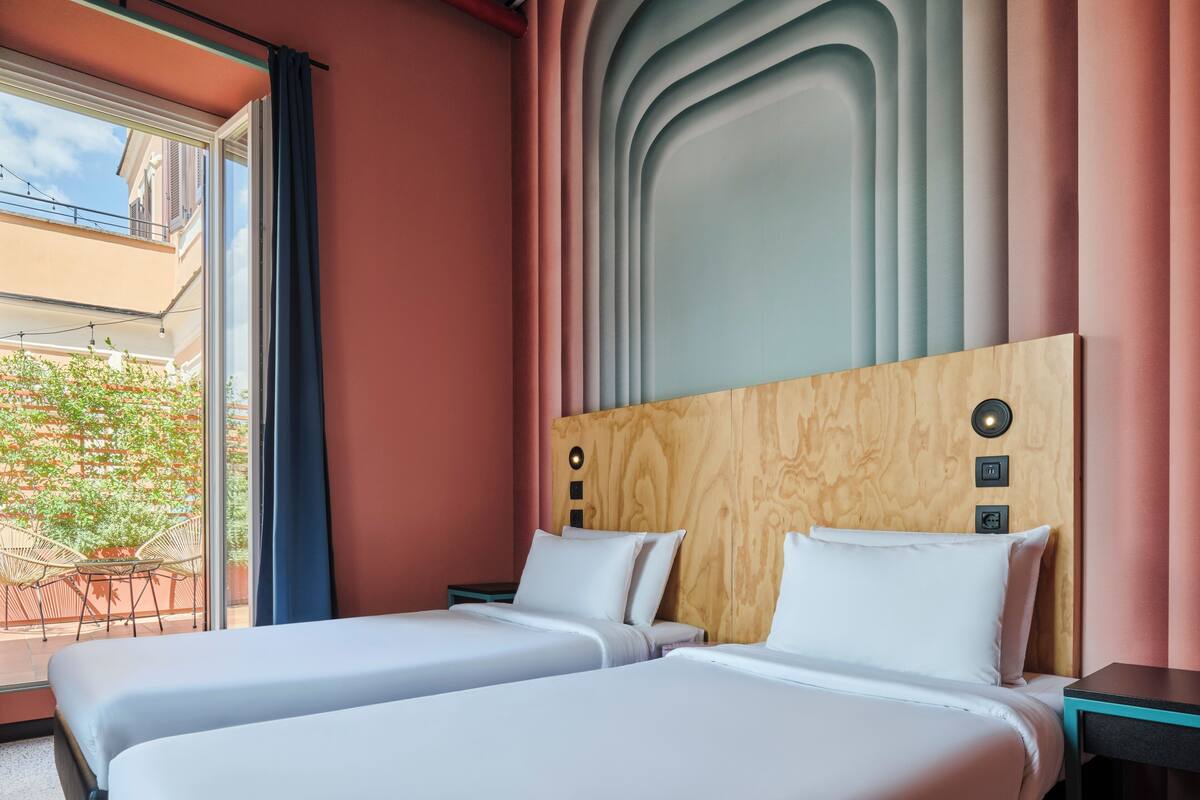Ein Hotelzimmer mit zwei Betten, einem Holzkopfteil, einem Fenster mit Vorhängen und Blick ins Freie.