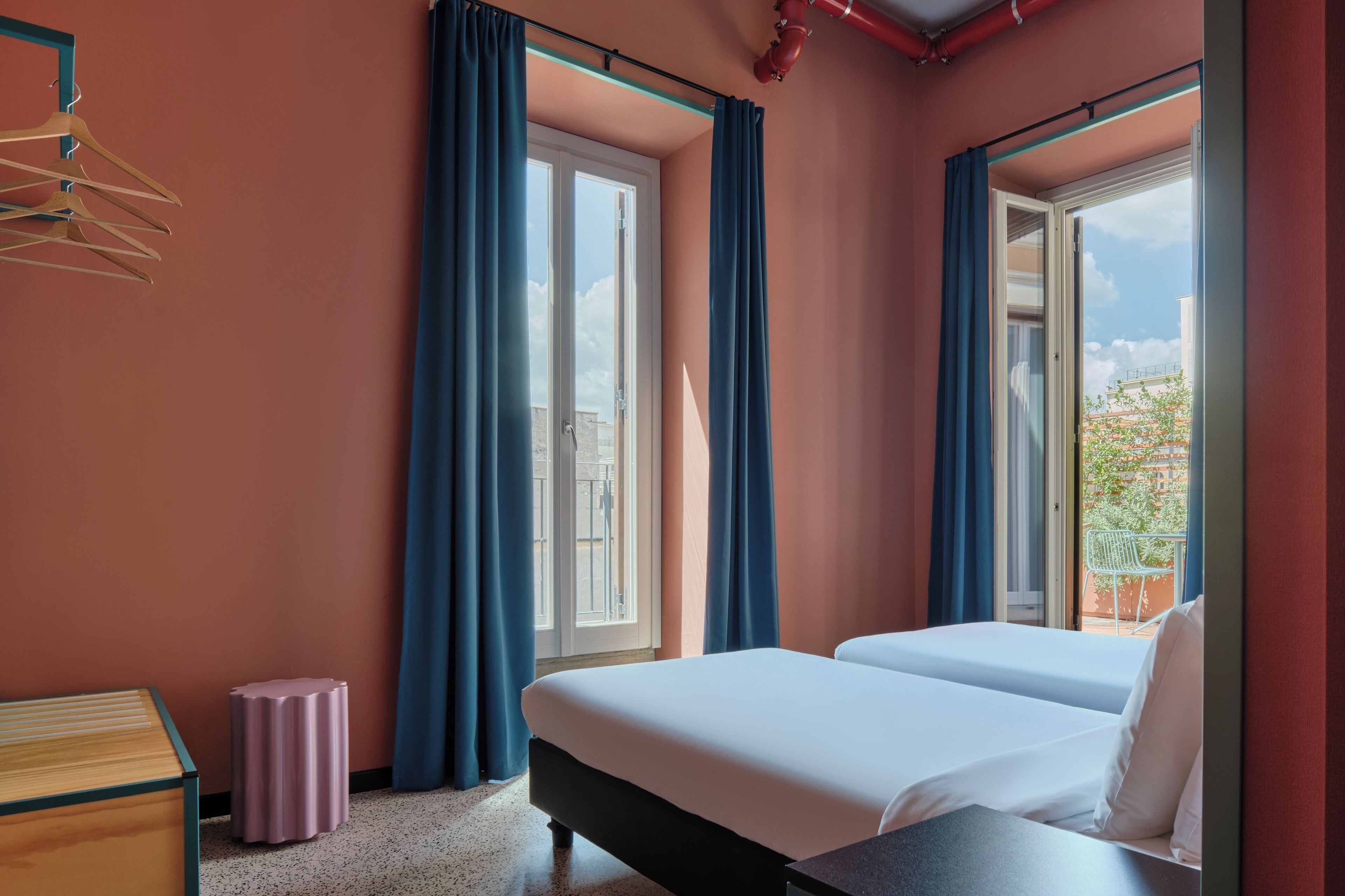 Een hotelkamer met twee bedden, een kleine tafel, een kruk en een raam met gordijnen.