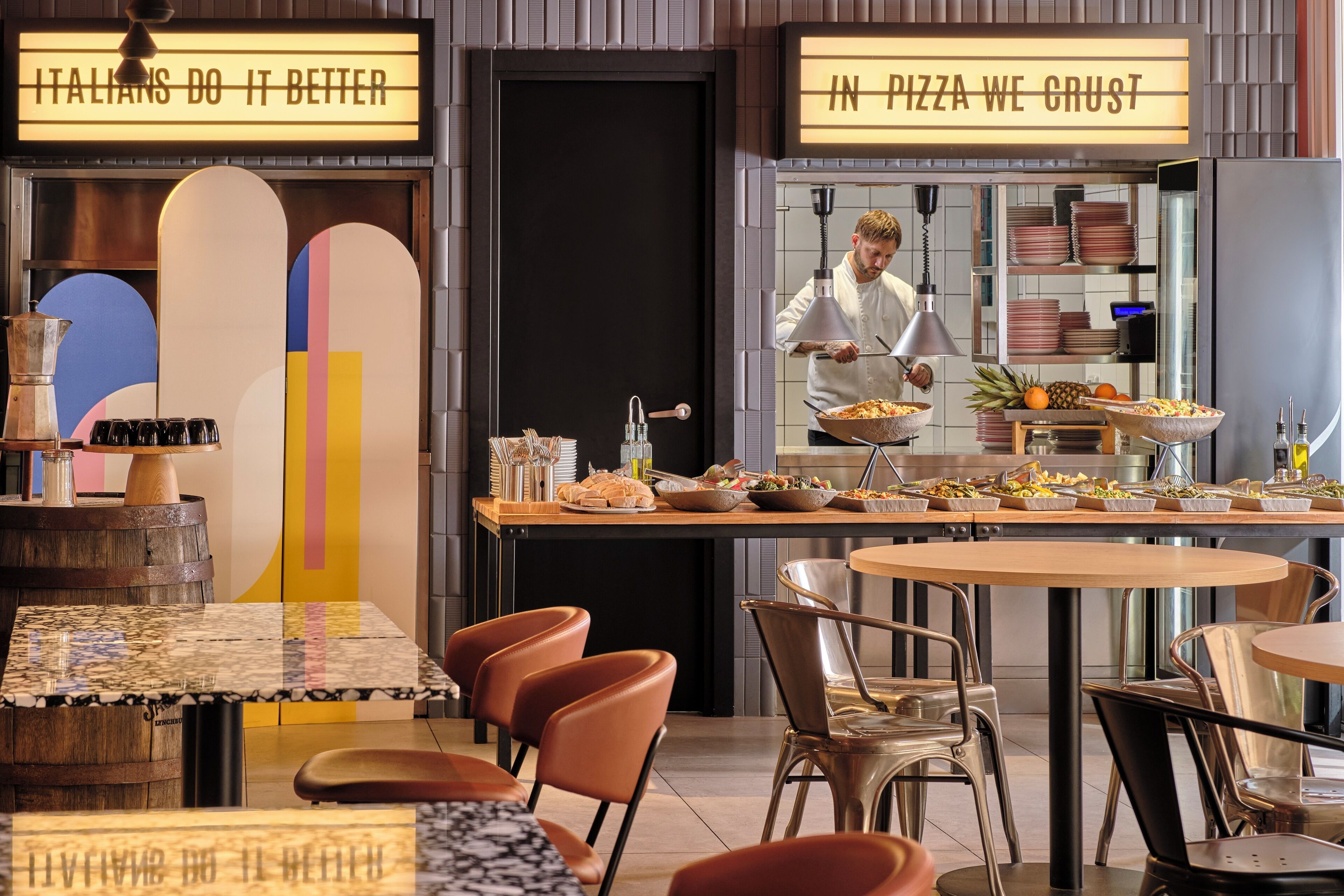 A restaurant interior with a buffet setup, a chef preparing food, and a sign that reads 'IN PIZZA WE CRUST'.