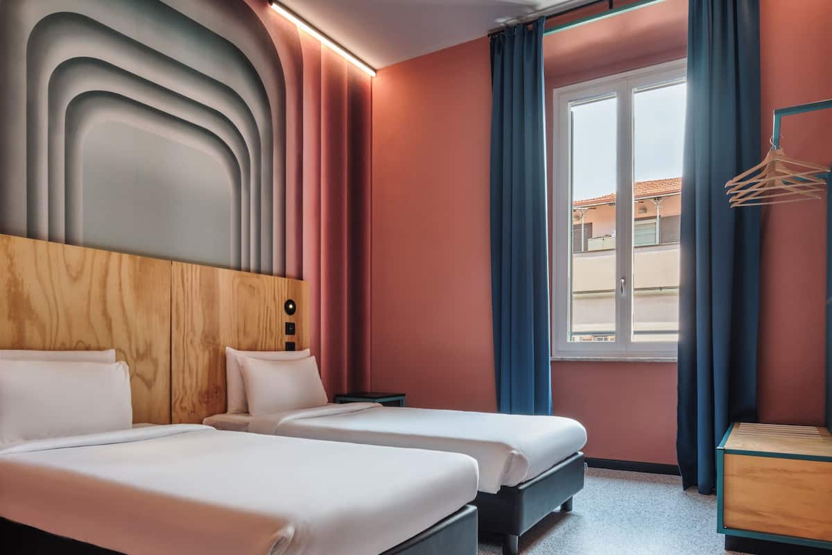 Ein Hotelzimmer mit zwei Betten, einem Holz-Kopfteil, einem kleinen Nachttisch und einem Fenster mit Vorhängen.