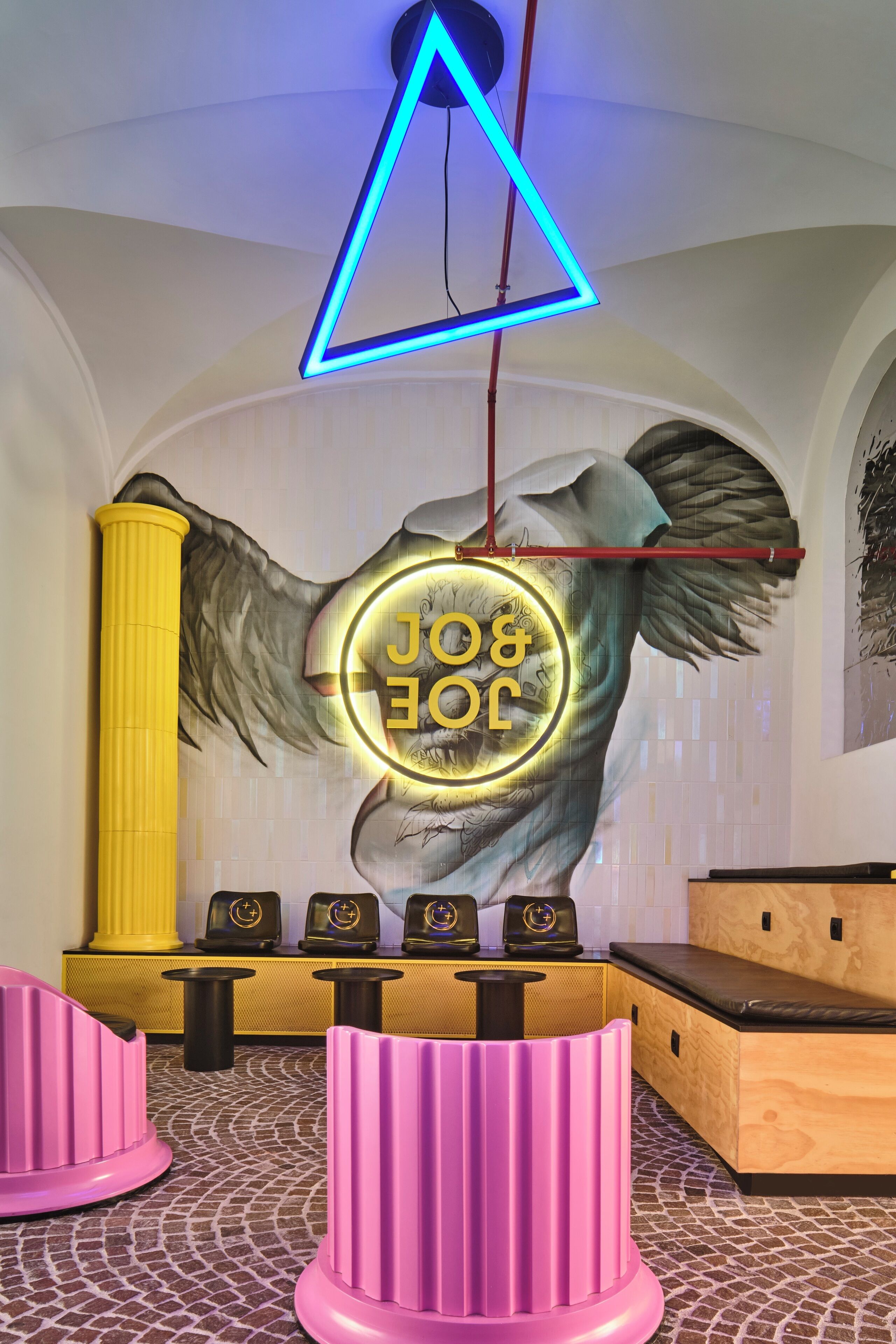 A room with a mural of an angel, a neon sign, a yellow pillar, a wooden bench, and a pink pillar.