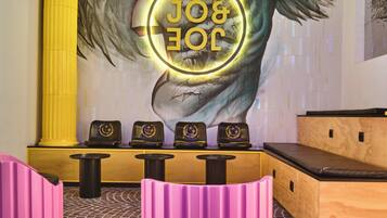A room with a mural of an angel, a neon sign, a yellow pillar, a wooden bench, and a pink pillar.