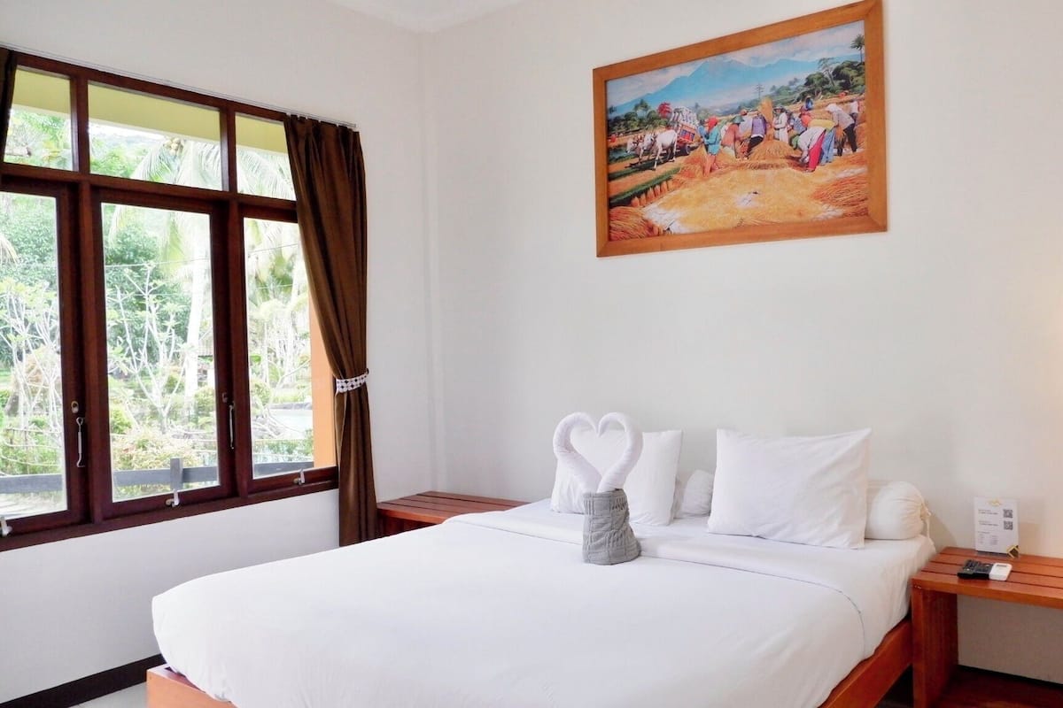 Een hotelkamer met een bed, nachtkastje, lamp, schilderij en raam met gordijnen.