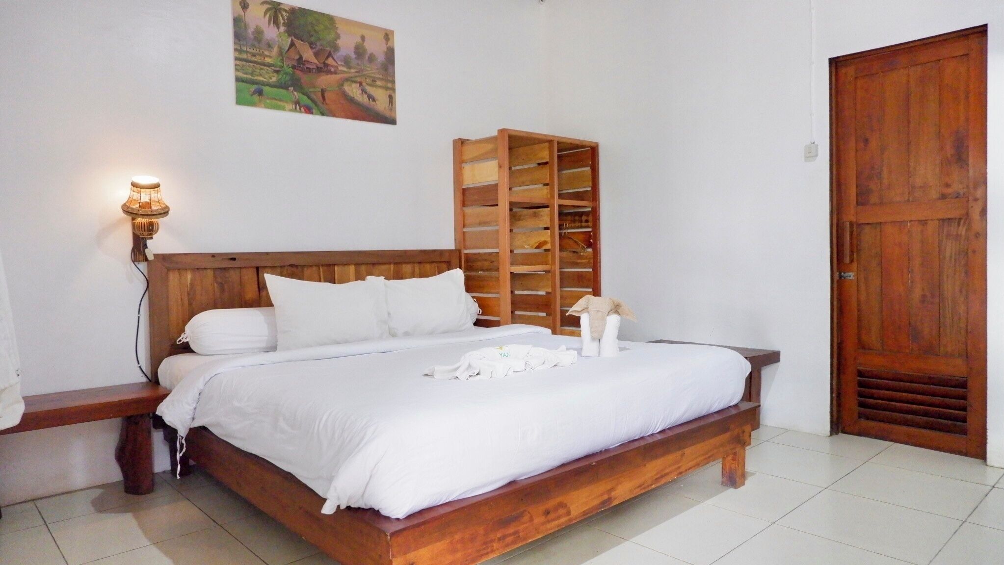 Quarto com cama de madeira, roupa de cama branca, mesa de cabeceira com abajur, guarda-roupa de madeira e uma porta.