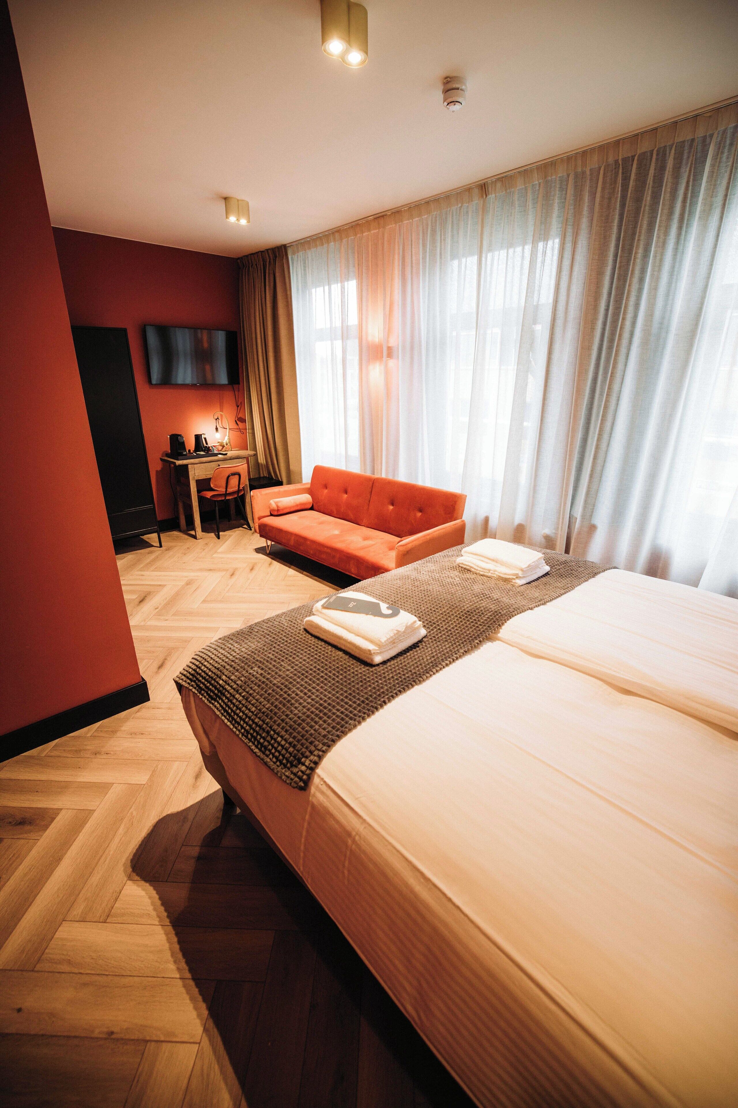 A hotel room with a large bed, a sofa, a desk with a chair, and a TV mounted on the wall.