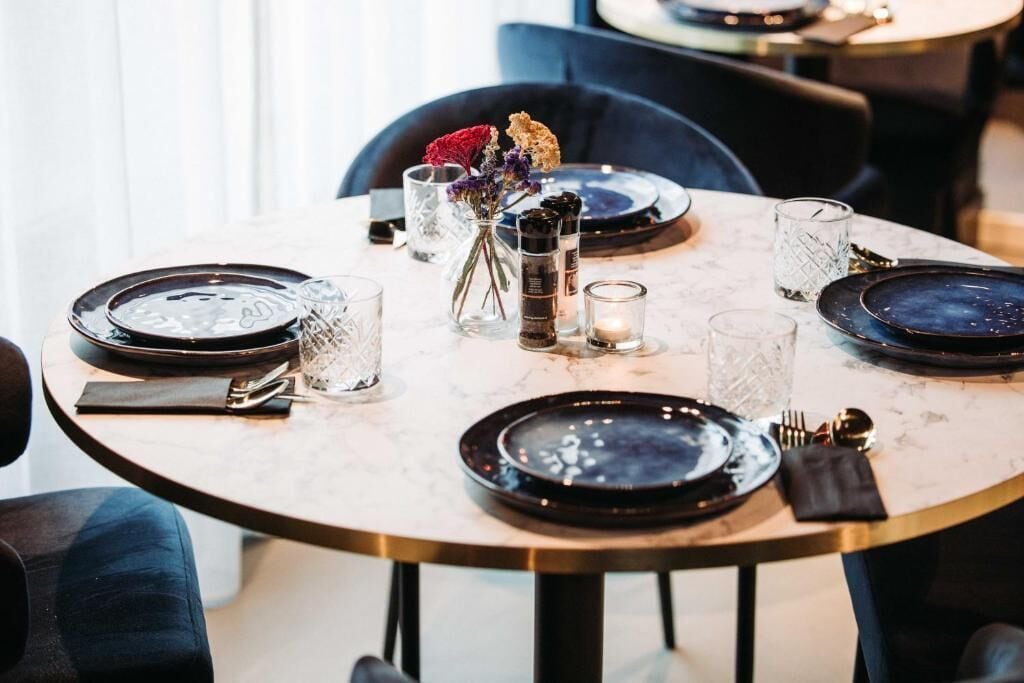 Una mesa de comedor dispuesta con platos negros, vasos transparentes y un florero con flores secas.