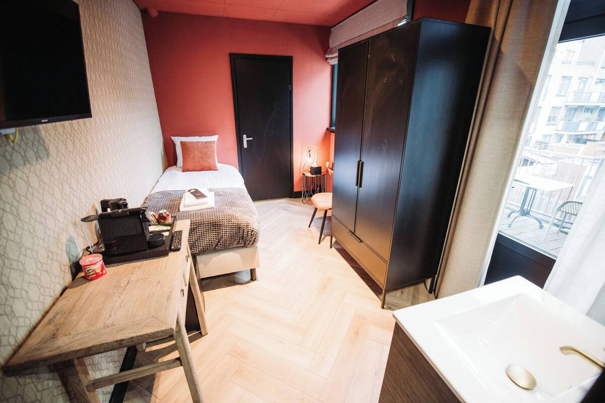 Una habitación de hotel compacta con una cama, un escritorio con una computadora portátil, una mesita con un teléfono y una taza, un armario y una ventana con vistas a edificios.
