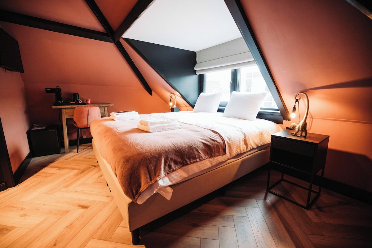 Un dormitorio moderno con techo inclinado, piso de madera, cama con ropa blanca, mesita de noche con lámpara y escritorio con silla.