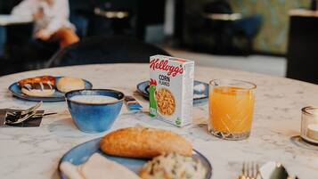 Un desayuno con un bol de cereal, un vaso de zumo de naranja y una hogaza de pan sobre una mesa de mármol.