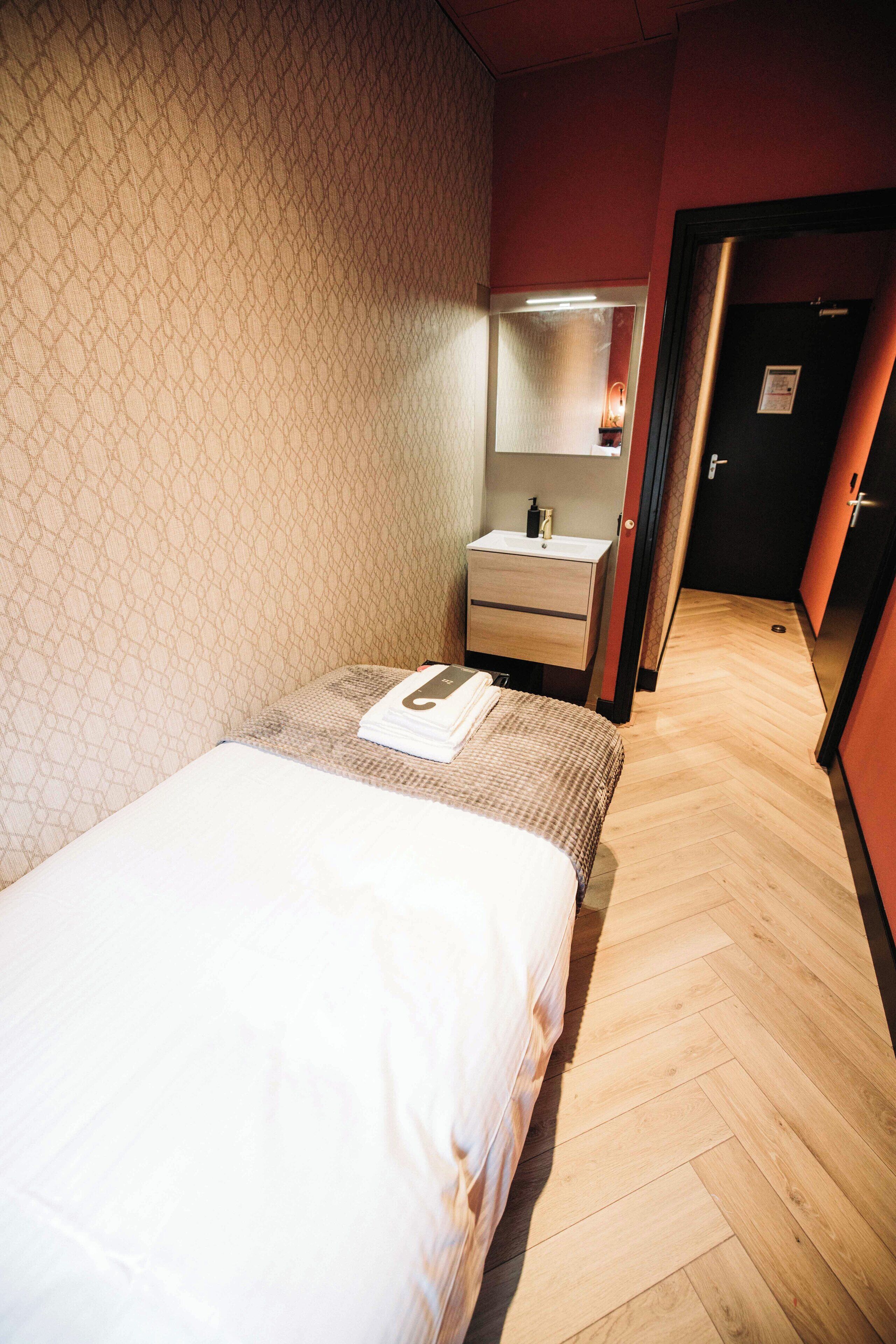 Une chambre d’hôtel avec un lit, une petite table de chevet, un miroir et un parquet en bois disposé en motif à chevrons.