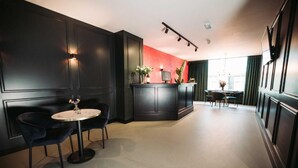 A modern interior with a dark wood reception desk, a round table with two chairs, and a seating area with a dark-colored sofa and a small round table. - Boutique Hotel Lupo  (Vlissingen)