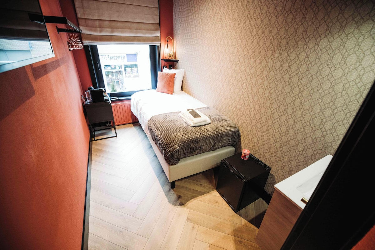 Un dormitorio con cama, mesita de noche y ventana con vistas a edificios.