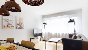 A modern living room with a wooden dining table, a sofa, and a TV. There are two pendant lights and a floor lamp. - Nest - Riehenring (Basel)