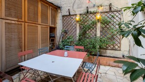 Une terrasse avec une table blanche et des chaises rouges et bleues, entourée de volets en bois et de panneaux ajourés décorés de guirlandes lumineuses.