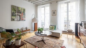 Un salon moderne avec un canapé, une table basse et des plantes en pot. De grandes fenêtres sont ornées de rideaux légers, et on y trouve également une lampe sur pied et une bibliothèque.