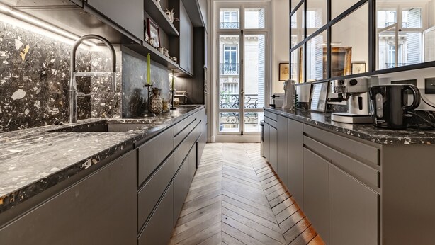 A modern kitchen with dark cabinetry, a herringbone floor, and a view of a balcony through a glass door. - Avenue Heights, by Veeve (Paris)