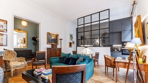 A living room with a sofa, chairs, a coffee table, and a dining table. There is a kitchen area with cabinets and a window. - Avenue Heights, by Veeve (Paris)