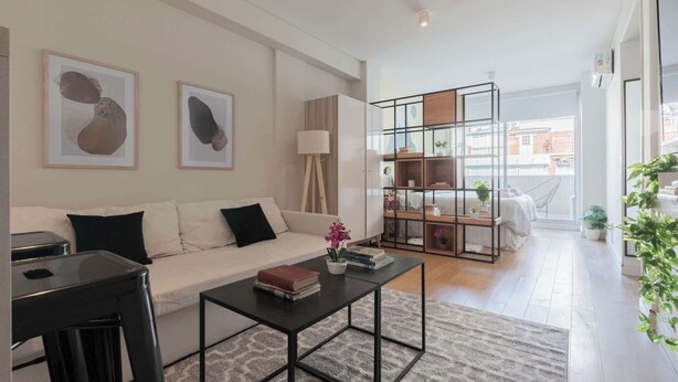 Ein modernes Wohnzimmer mit einem weißen Sofa, schwarzen Kissen, einem schwarzen Couchtisch und abstrakter Kunst an der Wand. Eine Glastrennwand beherbergt Bücher und Pflanzen, und große Fenster bieten einen Blick ins Freie.