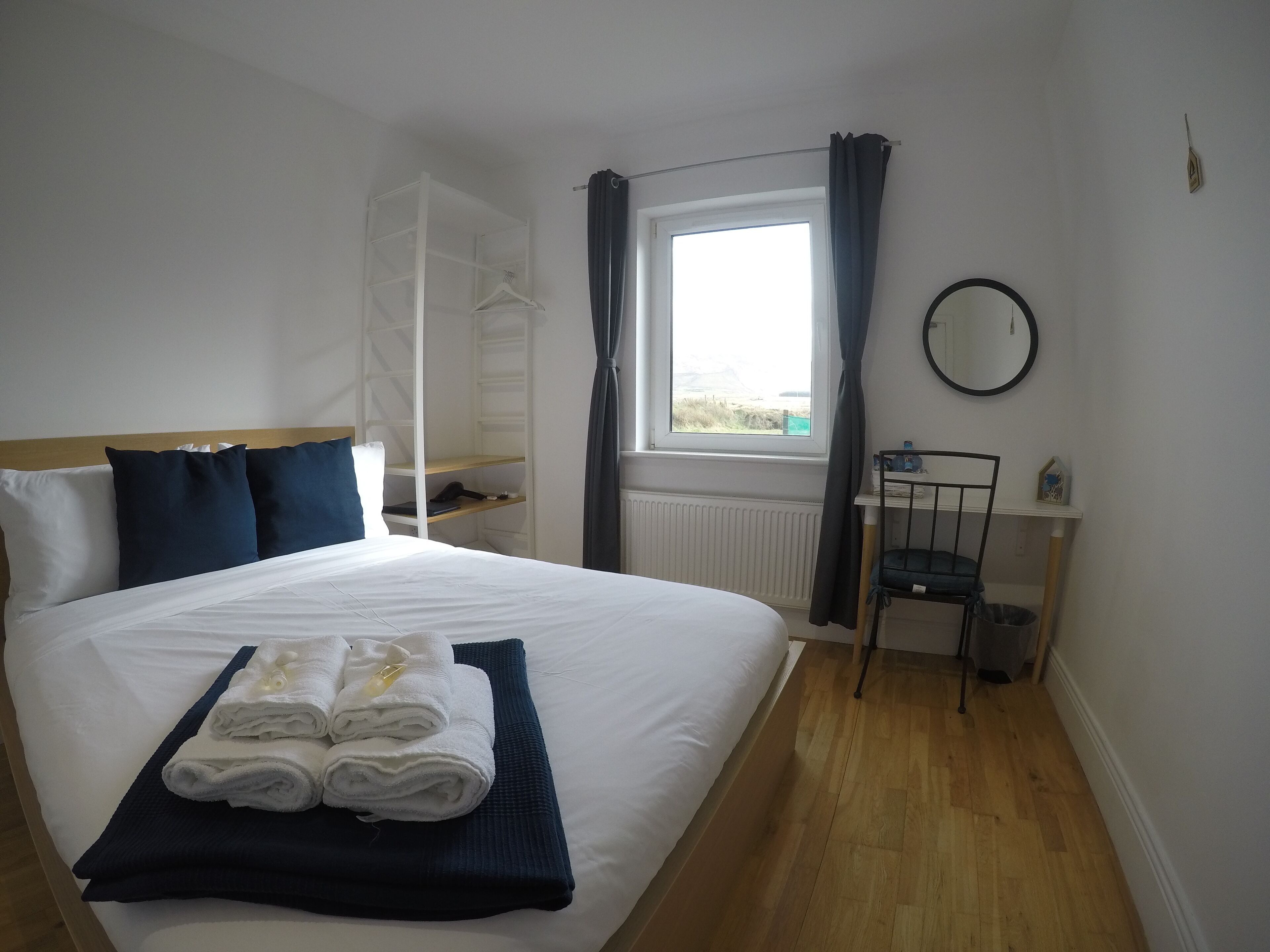 A neatly made bed with white and navy bedding, a set of folded towels, a white wardrobe, a window with curtains, a round mirror, and a small table with a chair.