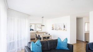 Una sala de estar moderna con un sofá negro, cojines azules, una zona de comedor con una mesa y sillas, y una cocina al fondo.