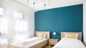 Un dormitorio con dos camas individuales, una mesita de noche con lámpara y cortinas traslúcidas en la ventana.