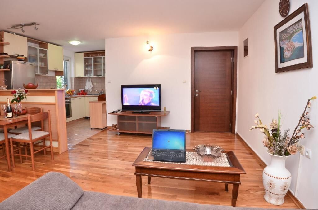 Ein Wohnzimmer mit Holzboden, einem Essbereich mit Tisch und Stühlen, einer Küche im Hintergrund, einem Fernseher auf einem Schrank und einer Tür.