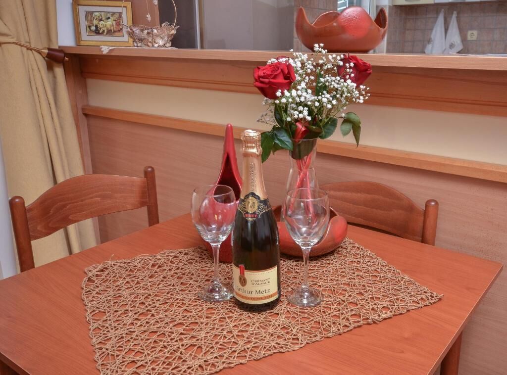 Una mesa de madera con un mantel tejido, una botella de vino y dos copas. Un jarrón con rosas rojas y espirales adorna la mesa.