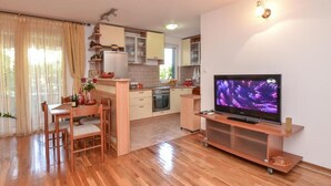 A modern kitchen with wooden cabinets, a dining area with a table and chairs, and a flat-screen TV on a wooden stand. - Apartments Petkovic Budva (Budva)