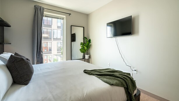 A hotel room with a bed, a TV mounted on the wall, a window with curtains, and a plant. - Luxury Downtown Condo (Columbus)