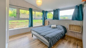 Une chambre à coucher avec un lit, de grandes fenêtres avec des rideaux, un luminaire suspendu et une vue sur la verdure à l’extérieur.