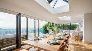 A modern dining area with a wooden table set for six, large windows, and a view of greenery.