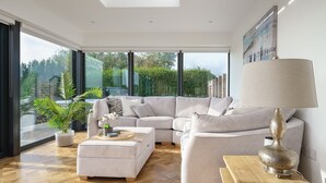 A modern living room with a sectional sofa, a coffee table, and a lamp. There is a large window providing a view of greenery outside.