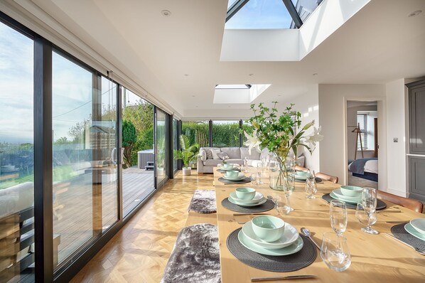 A modern dining area with a long table set for a meal, large windows offering a view of the outdoors, and a cozy seating area with a sofa and cushions. - Groeslon -  Luxury cottages (Menai Bridge)