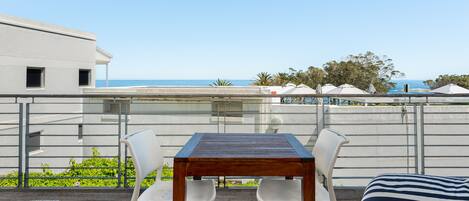 A terrace with a wooden table and white chairs, overlooking a beach and palm trees.